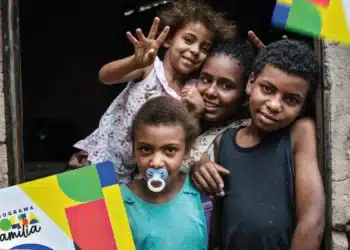 Família sorridente posando em frente à casa com cartões do programa Bolsa Família nas mãos.