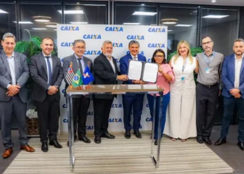 Está no CadÚnico? Veja como acessar o novo microcrédito de até R$ 21 mil 5 Grupo de pessoas formalmente vestidas segura contrato assinado em mesa com bandeiras, painel CAIXA ao fundo.