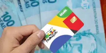 Cartão do Bolsa Família segurado por uma pessoa, com notas de real ao fundo, representando o pagamento antecipado do benefício em cidades de Minas Gerais.