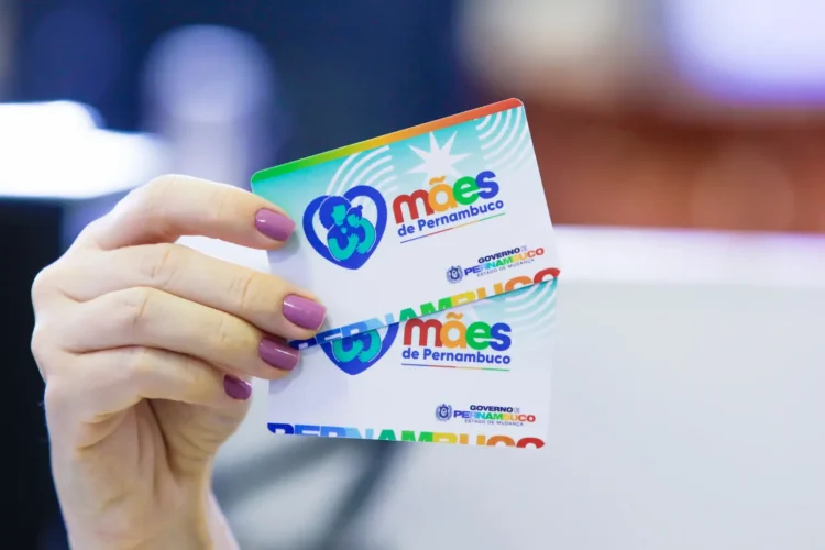 Mão feminina com unhas pintadas segura dois cartões do programa Mães de Pernambuco em destaque.