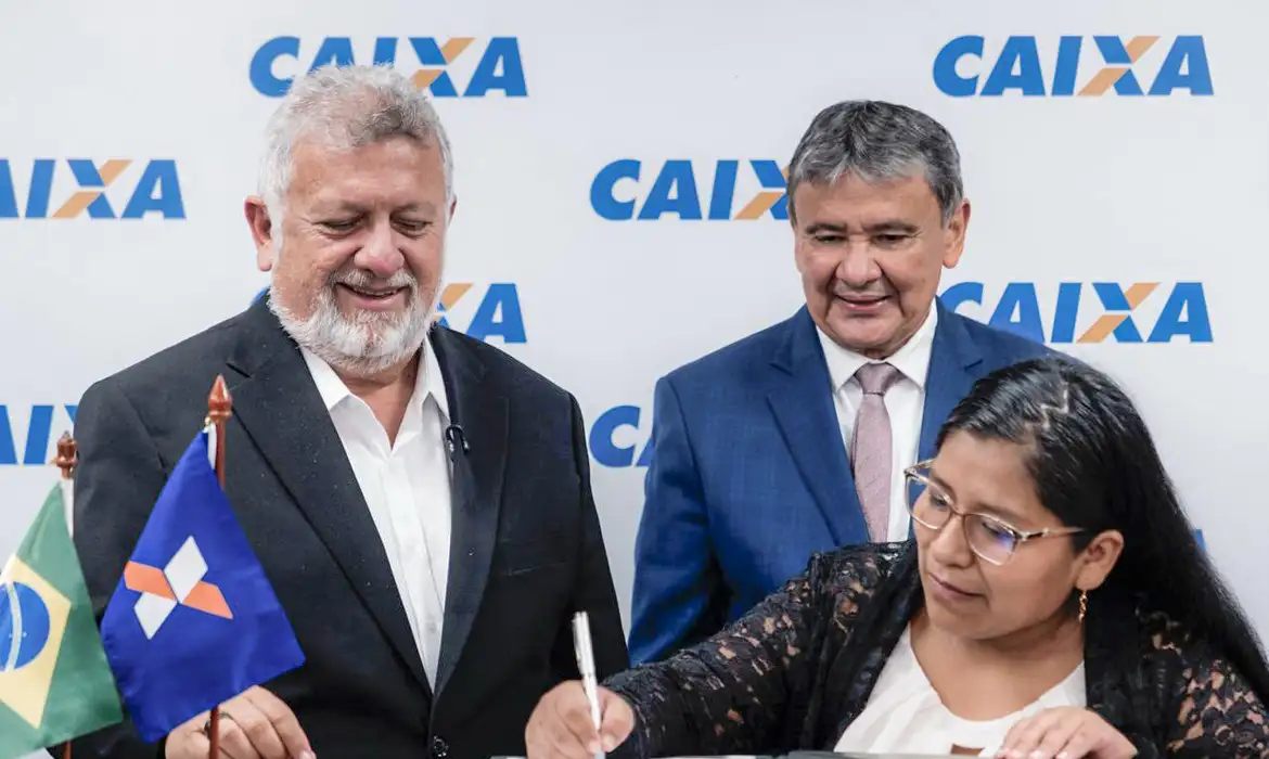 Assinatura de contrato de microcrédito com participação da Caixa em 2026. Mulher assina documento em mesa acompanhada por dois homens sorridentes ao fundo, com logotipo da Caixa.