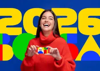 Bolsa Família: valor médio chega a R$ 690,01 por família em fevereiro 7 Mulher com suéter vermelho segura cartão do Bolsa Família sorrindo em frente a painel colorido com 2026.