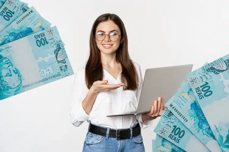 Imposto de Renda 2026 Mulher sorrindo segura notebook e aponta para notas de 100 reais espalhadas